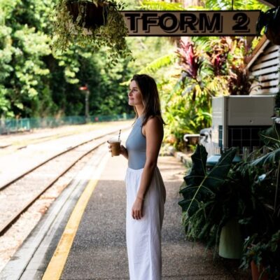 Young woman stands on the rainforest surrounded Kuranda Scenic Railway platform holding an iced coffee.
